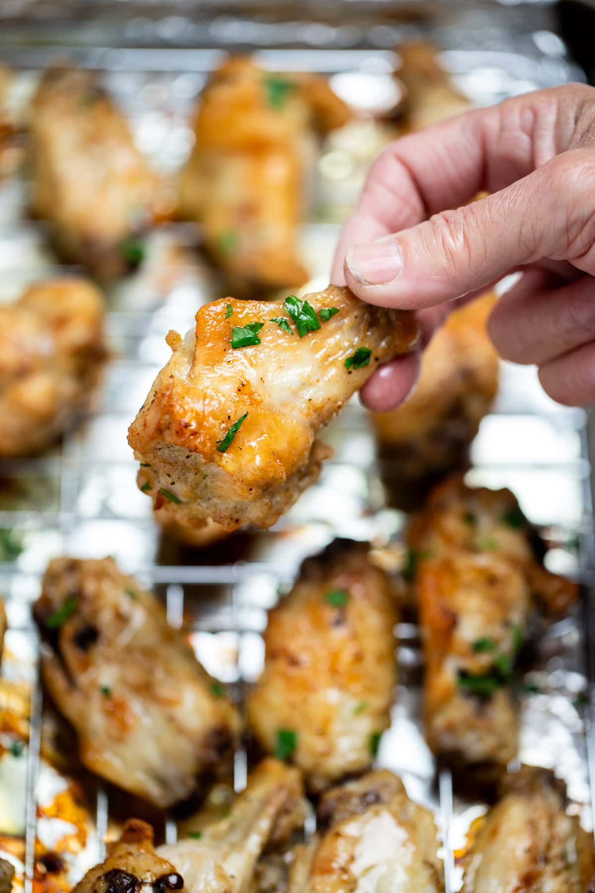 A hand holding a chicken wing above a baking rack of cooked chicken wings topped with fresh parsley.