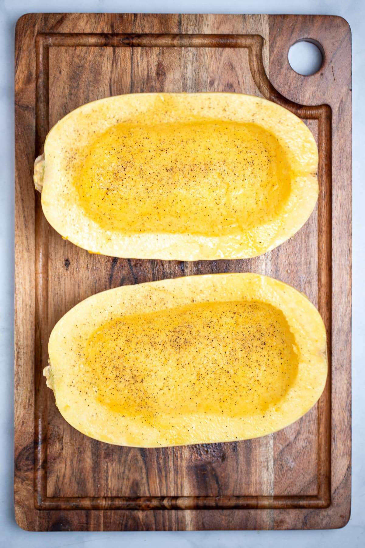 Two halves of a spaghetti squash open-side-up on a cutting board with seasoning, before being roasted.