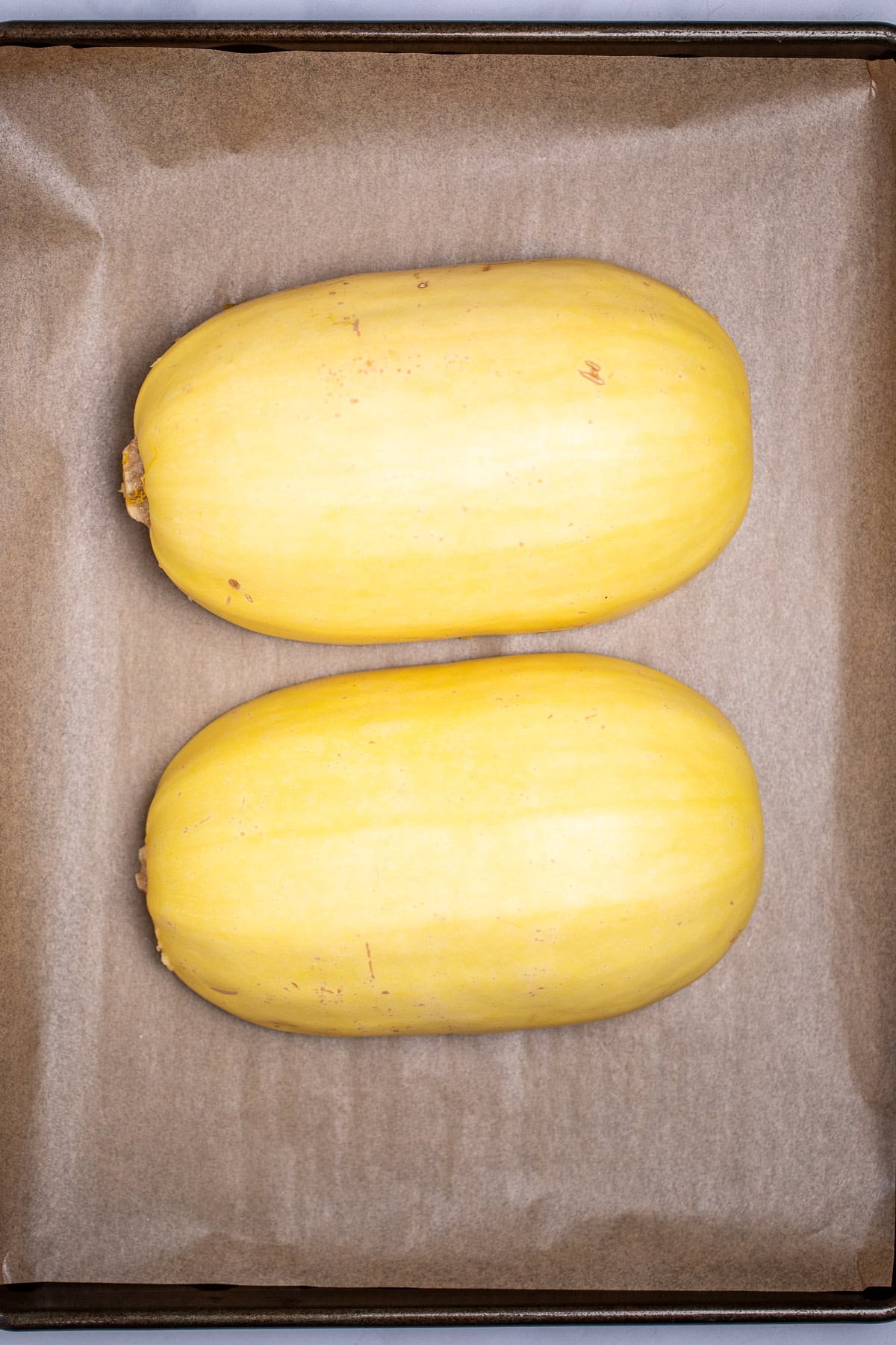 Two halves of a spaghetti squash open-side-down on a sheet pan lined with parchment paper before being baked.