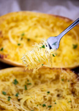 A fork with spaghetti squash noodles being held above two halves of cooked spaghetti squash, topped with fresh chopped parsley.