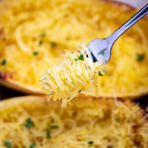 A fork with spaghetti squash noodles being held above two halves of cooked spaghetti squash, topped with fresh chopped parsley.