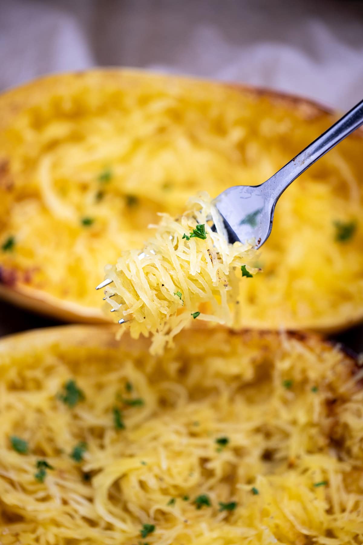 A fork with spaghetti squash noodles being held above two halves of cooked spaghetti squash, topped with fresh chopped parsley.