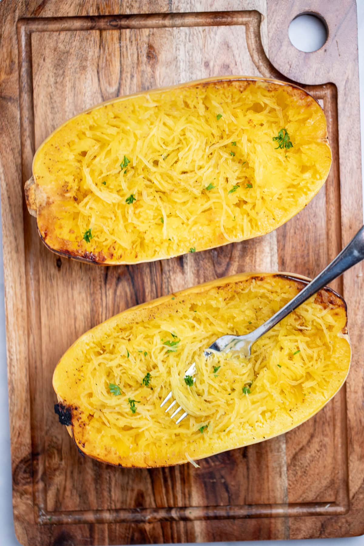 A fork with spaghetti squash strands resting on top of one half of a spaghetti squash, next to the other half, sitting on a cutting board and topped with fresh chopped parsley.