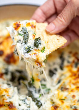 A dutch oven full of spinach artichoke dip with a caramelized top, topped with chopped green onions, with a hand holding a tortilla chip scooping up some dip.