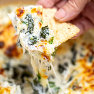 A dutch oven full of spinach artichoke dip with a caramelized top, topped with chopped green onions, with a hand holding a tortilla chip scooping up some dip.