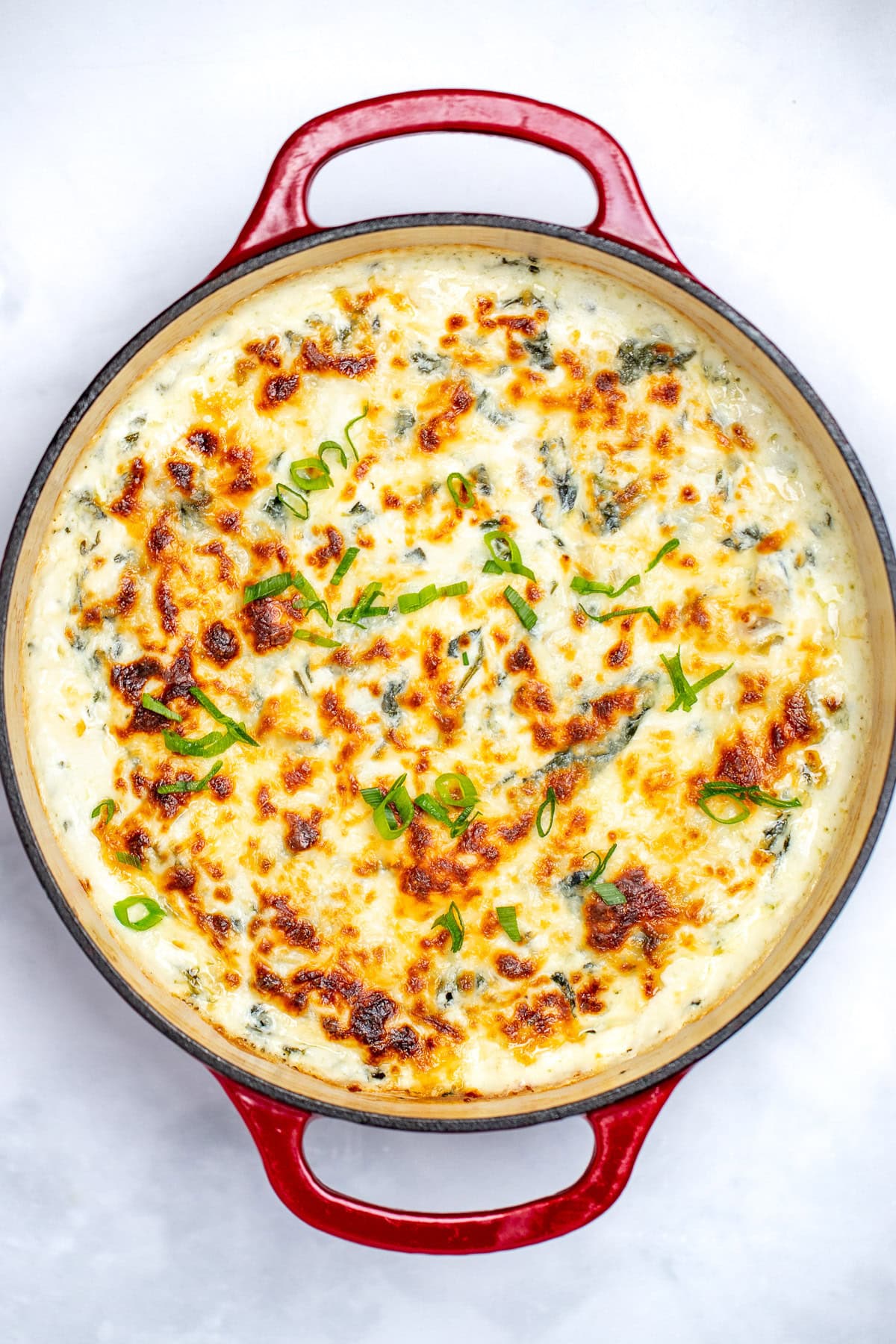 A dutch oven full of spinach artichoke dip with a caramelized top, topped with chopped green onions.