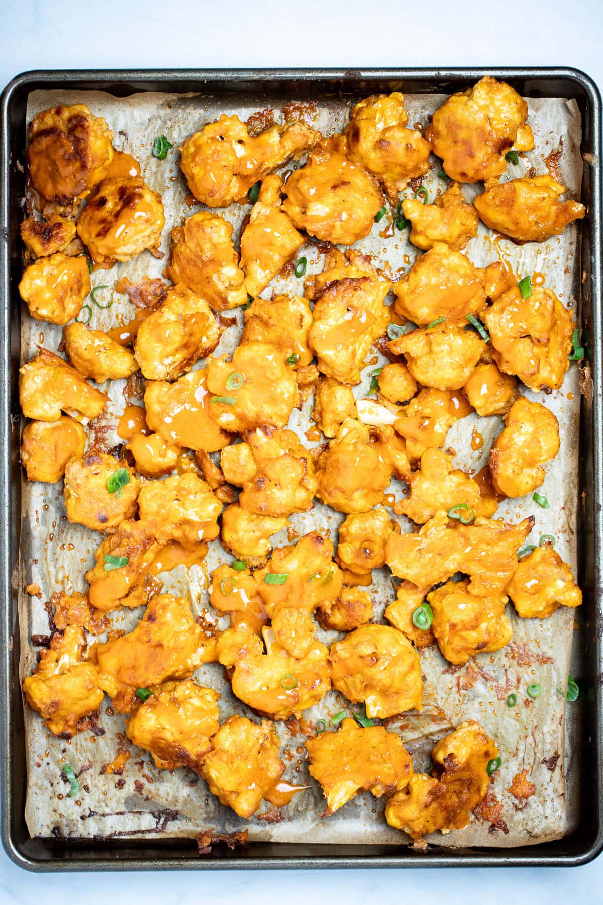 Buffalo Cauliflower Wings on a sheet pan lined with parchment paper, in a single layer, topped with extra buffalo sauce and green onions.