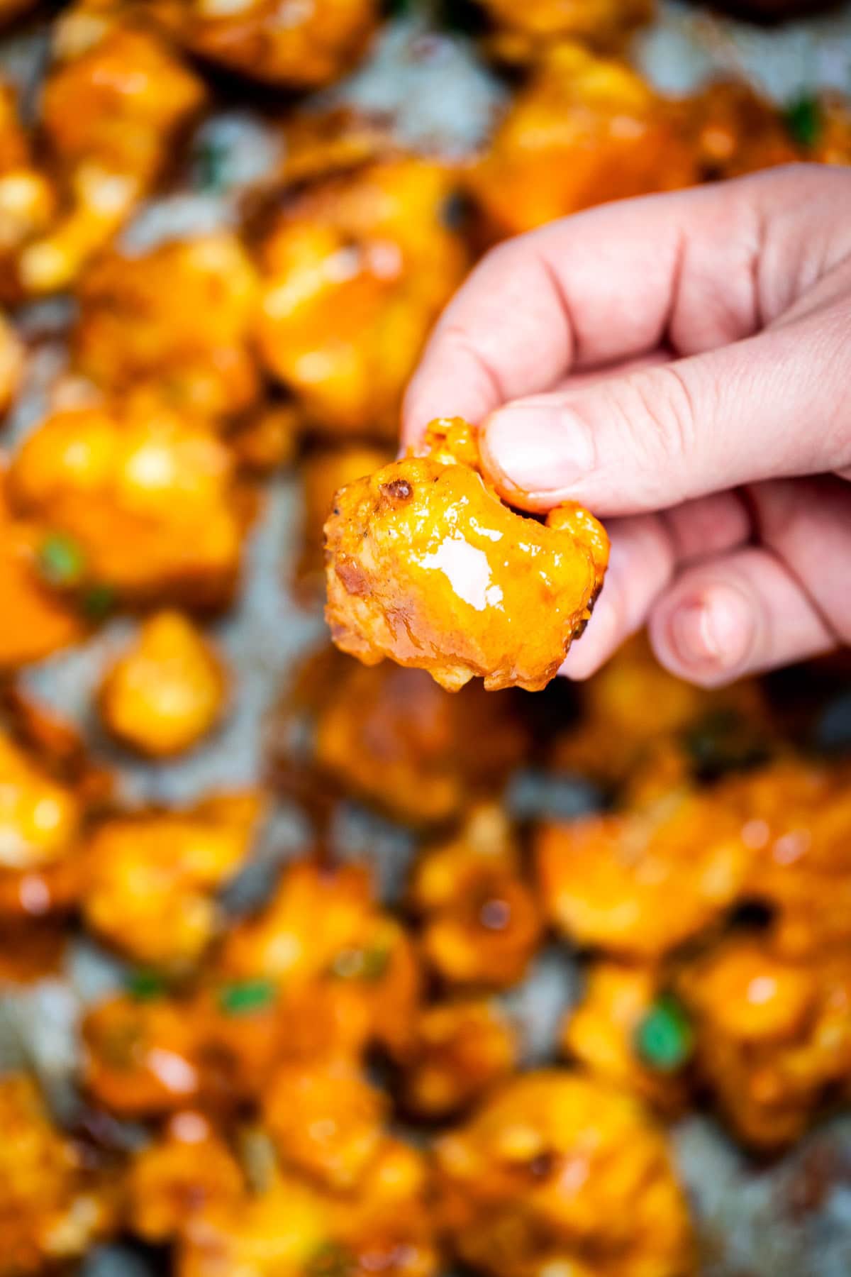 A hand holding a buffalo cauliflower wing over a sheet pan of more cauliflower wings coated in buffalo sauce.