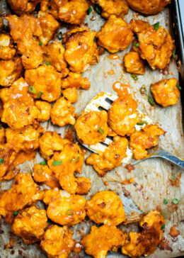 Buffalo Cauliflower Wings on a sheet pan lined with parchment paper and a spatula holding some cauliflower, resting on the sheet pan, topped with extra buffalo sauce and green onions.