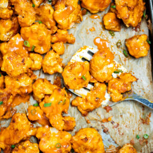 Buffalo Cauliflower Wings on a sheet pan lined with parchment paper and a spatula holding some cauliflower, resting on the sheet pan, topped with extra buffalo sauce and green onions.