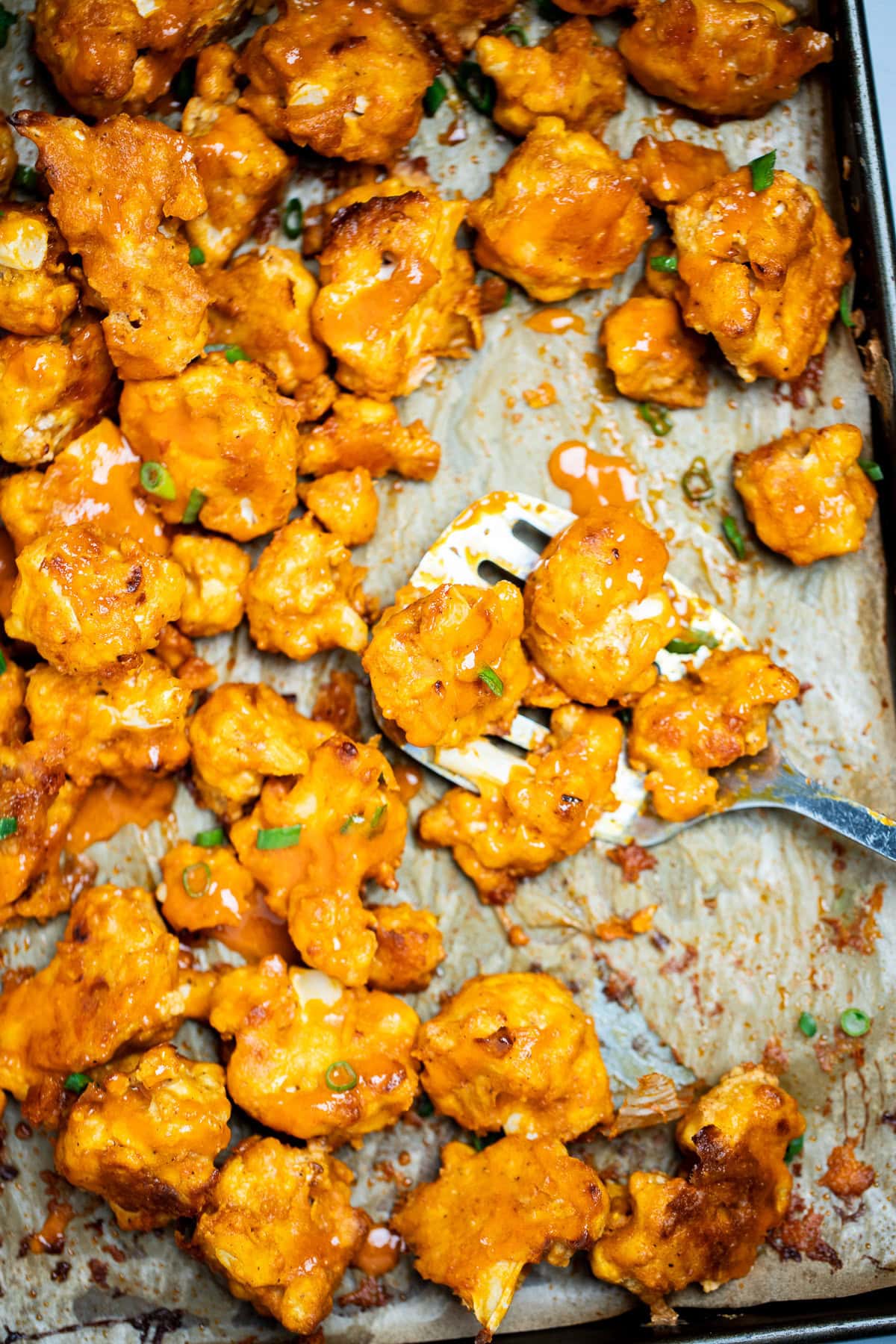 Buffalo Cauliflower Wings on a sheet pan lined with parchment paper and a spatula holding some cauliflower, resting on the sheet pan, topped with extra buffalo sauce and green onions.