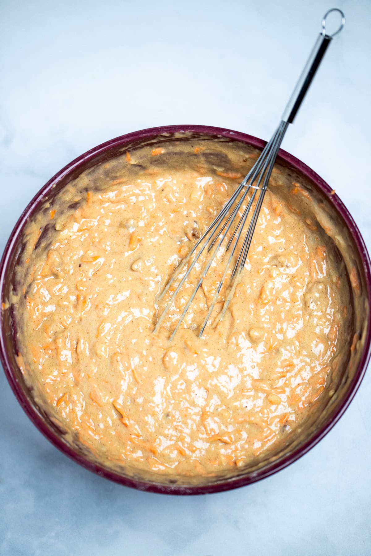 A mixing bowl on a table with carrot cake batter and a whisk in the batter.