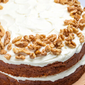 A double layer carrot cake on a wooden cake plate, with a layer of cream cheese frosting in the middle of the layers of cake, and on top, with walnuts around the edges of the top of the cake.