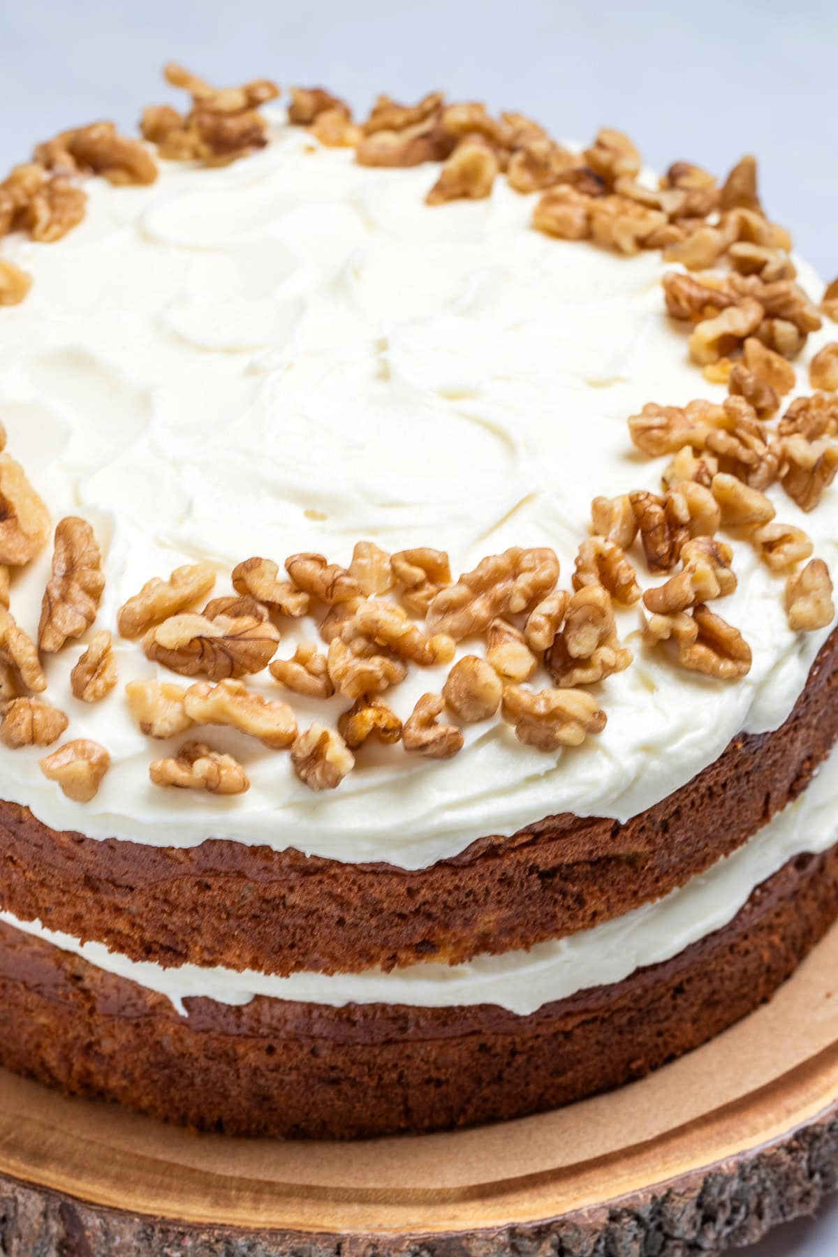 A double layer carrot cake on a wooden cake plate, with a layer of cream cheese frosting in the middle of the layers of cake, and on top, with walnuts around the edges of the top of the cake.