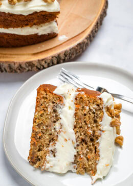 A piece of carrot cake on a plate with a fork, with a layer of cream cheese frosting in the middle and on top, with walnuts. There is a full cake in the background.