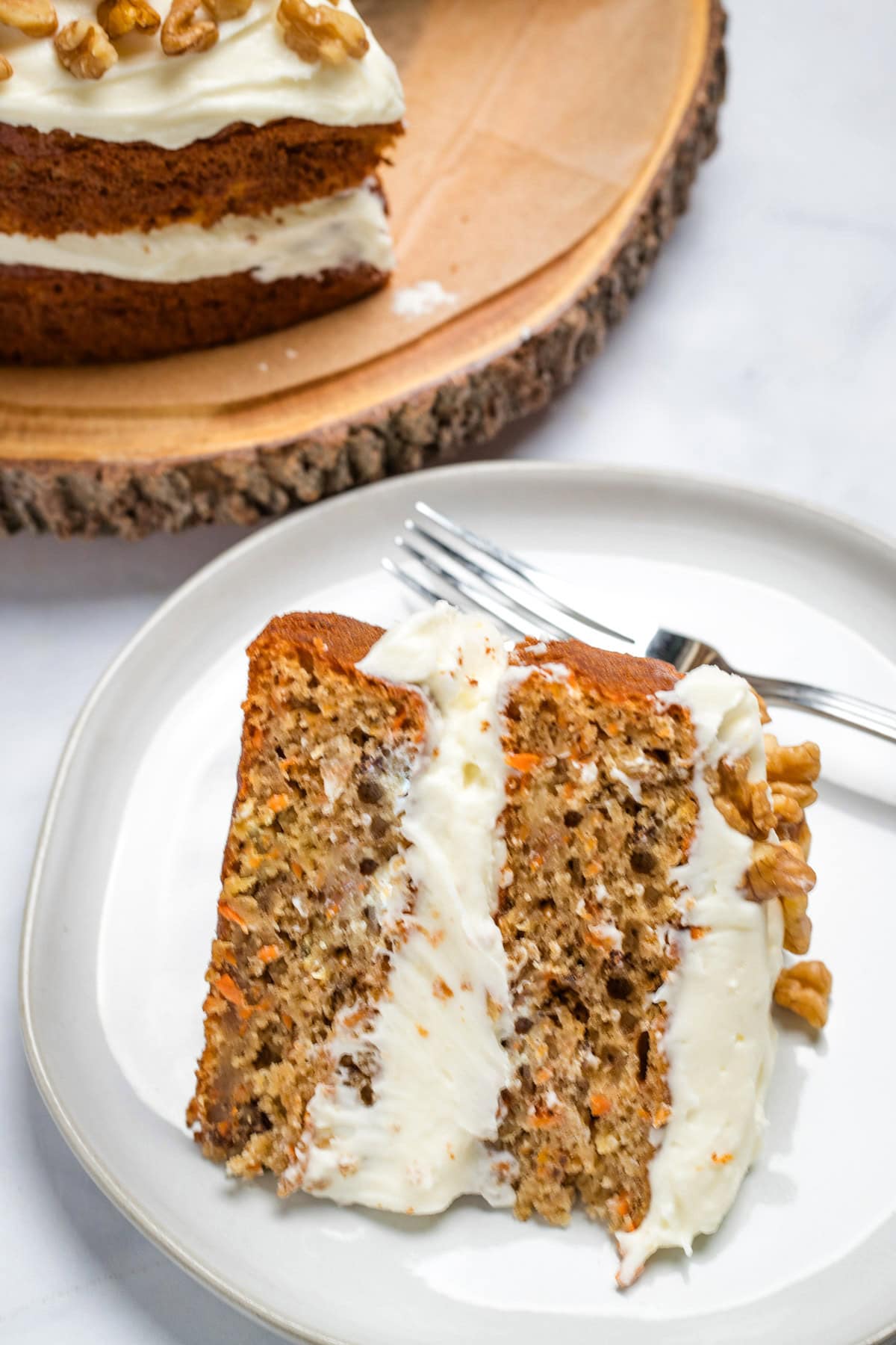 A piece of carrot cake on a plate with a fork, with a layer of cream cheese frosting in the middle and on top, with walnuts. There is a full cake in the background.