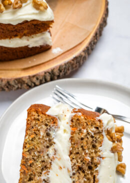 Pinterest pin with a piece carrot cake on plate with a fork, with a layer of cream cheese frosting in the middle and on top, with walnuts. There is a full cake in the background.