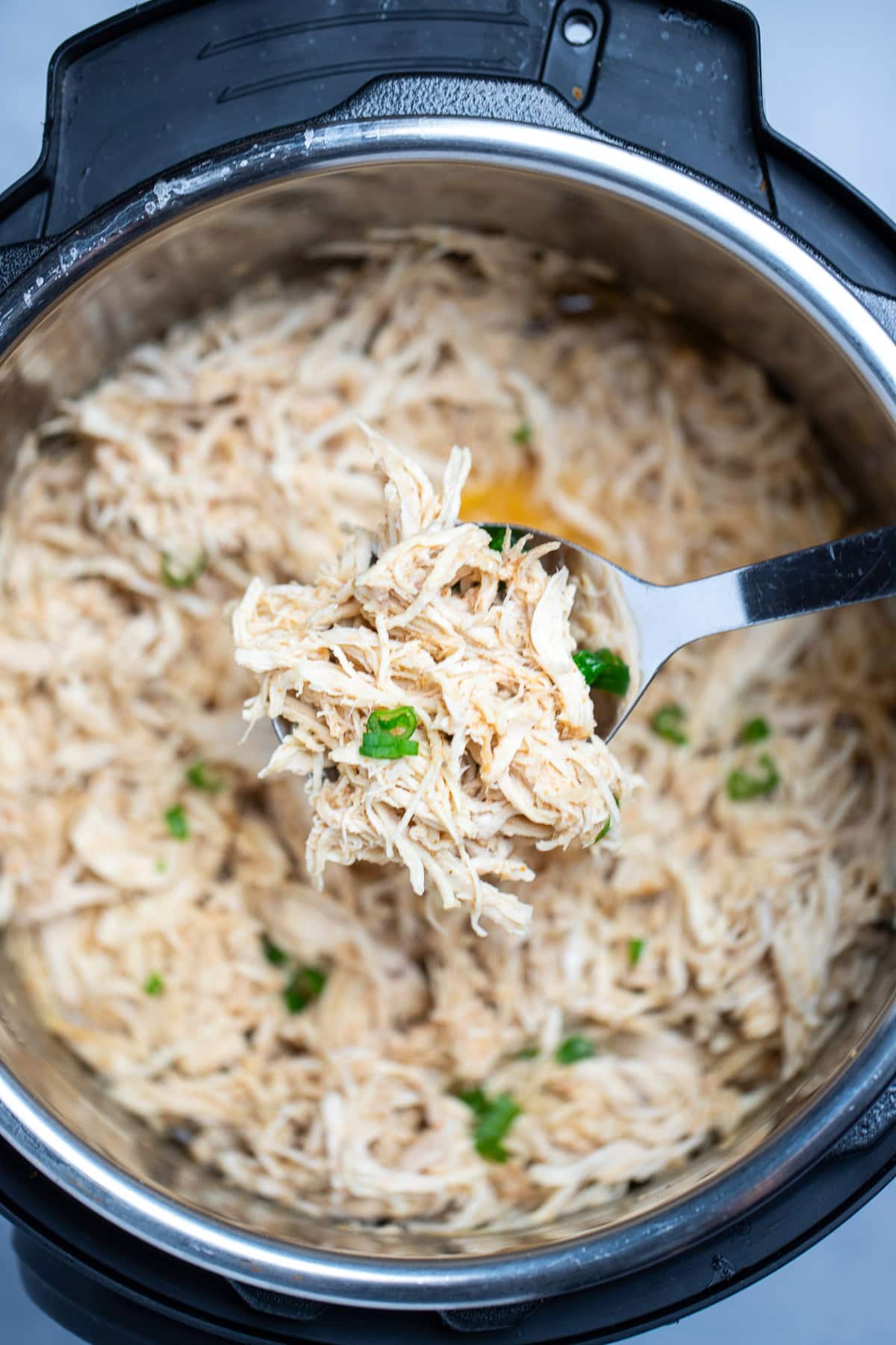 An instant pot with shredded chicken breast topped with fresh chopped green onions, with a spoon lifting some up above the pot.