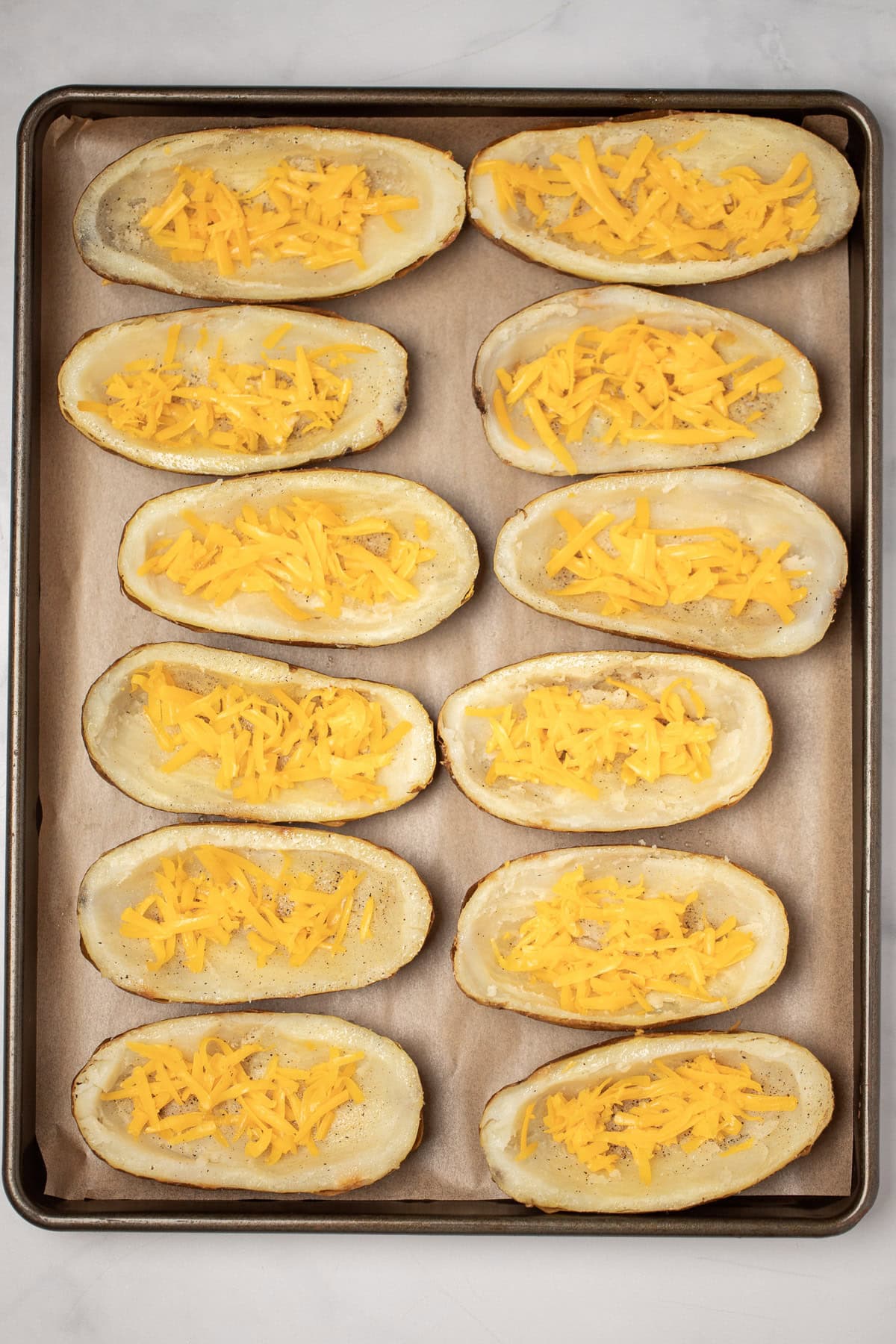 A sheet pan with parchment paper with baked potatoes cut in half, open side up, topped with spices and cheese before being baked.