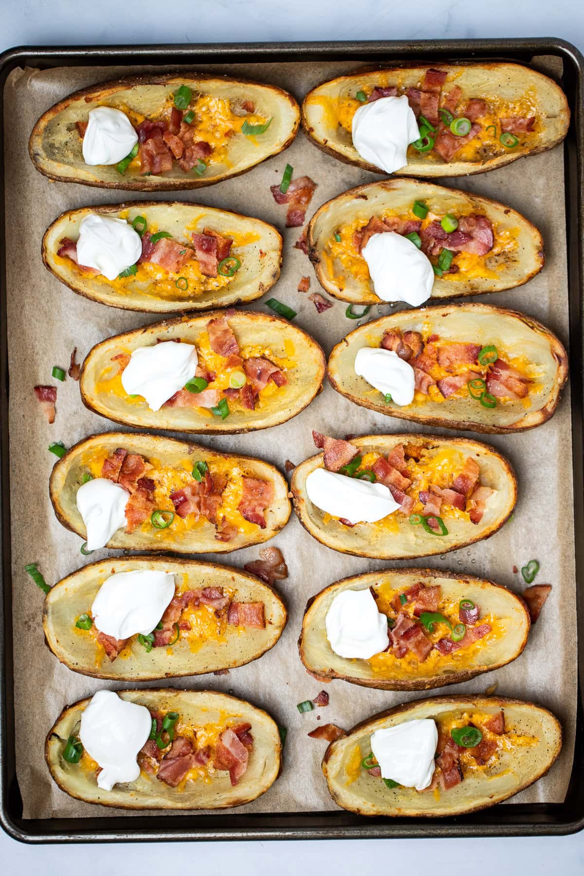 A sheet pan with parchment paper, with two rows of loaded potato skins topped with cheese, green onions, bacon, and sour cream.