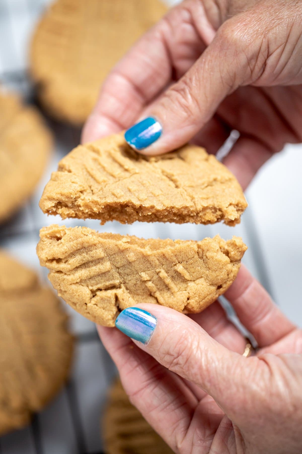 A hand breaking a gluten free peanut butter cookie in half, over a cooling rack of cookies.