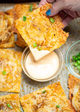 A hand dipping an onion ring chip into a small bowl of sriracha aioli, on a sheet pan of more chips topped with chopped green onion.