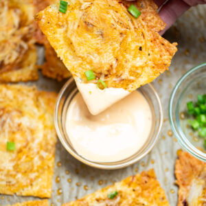 A hand dipping an onion ring chip into a small bowl of sriracha aioli, on a sheet pan of more chips topped with chopped green onion.