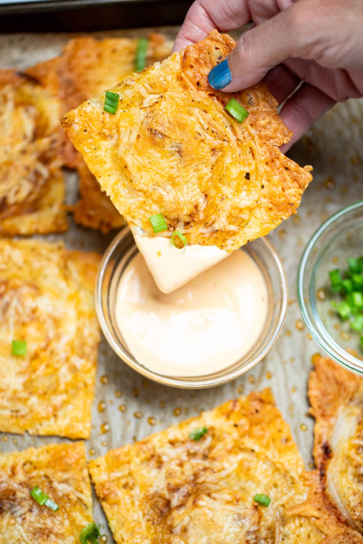 A hand dipping an onion ring chip into a small bowl of sriracha aioli, on a sheet pan of more chips topped with chopped green onion.