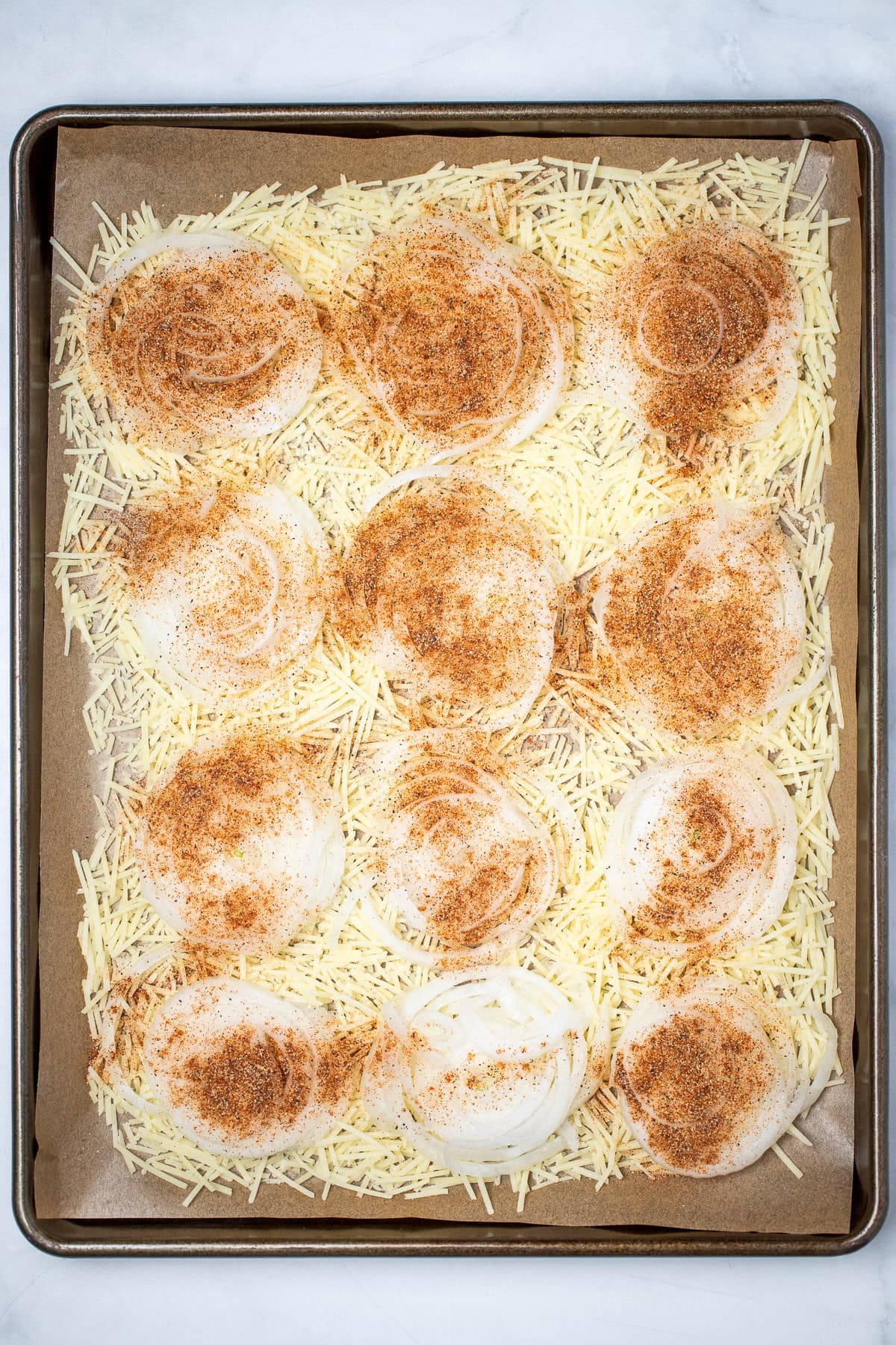 A sheet pan with parchment paper, with onion slices on top of shredded parmesan in a single layer, topped with seasoning before baking.