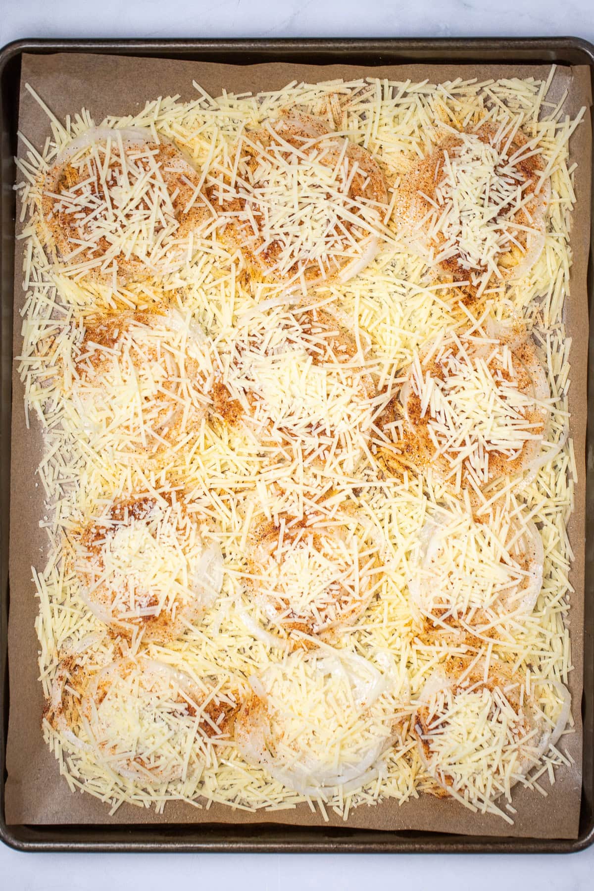 A sheet pan with parchment paper with a layer of shredded parmesan, then onion slices, then seasoning and olive oil, and a final layer of parmesan before baking.