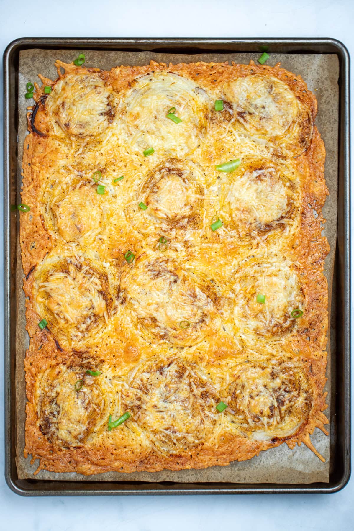 A parchment lined sheet pan with baked parmesan onion ring chips before being cut into pieces, topped with chopped green onion.