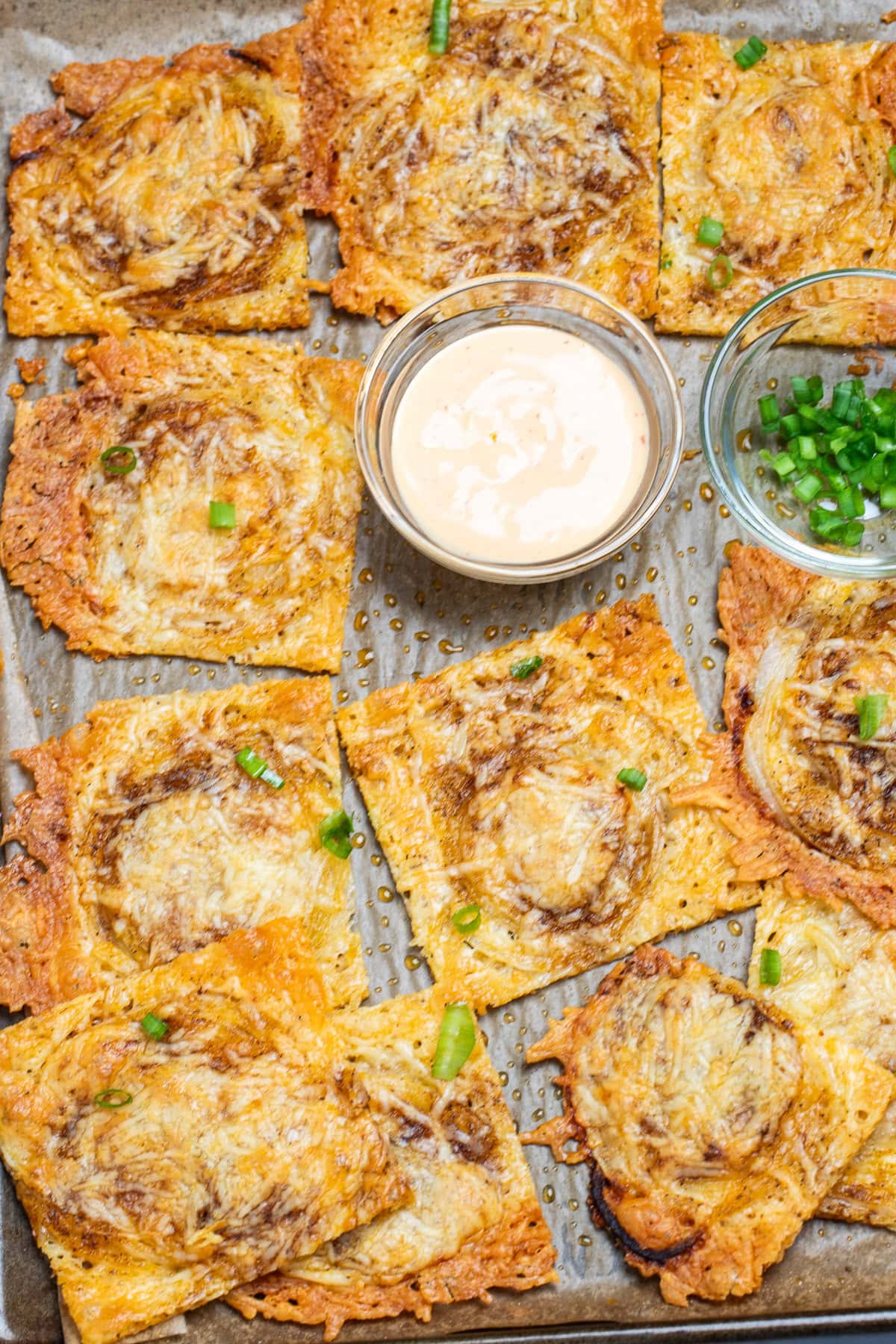 A sheet pan of parmesan onion ring chips, with a small bowl of sriracha aioli, topped with chopped green onions.